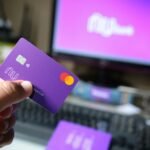 A close-up of a hand holding a purple bank card in front of a computer screen.